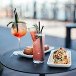Glasses with colorful cocktails garnished with green herbs and straws with watermelon slice near white plates with delicious appetizers on round table in street restaurant in daytime